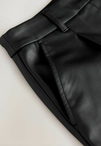 Nahaufnahme von schwarzen Kunstlederhosen, die den Bund, den Gürtelschlaufe, die Seitenasche und die strukturierte Oberfläche zeigen.