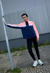 Chaqueta deportiva en azul marino y rosa con acentos en blanco, combinada con leggings negros y zapatillas verde claro. Tejido suave, cierre con cremallera, corte relajado.