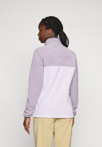 Kvinne med fletter iført en to-farget lilla fleecejakke og beige bukser, som står og vender bort fra en ensfarget bakgrunn.