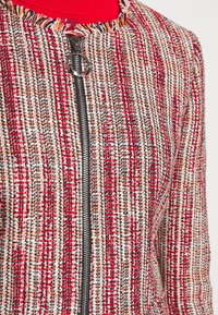 Rode en crème tweed jas met zwarte rits, ronde halslijn en franjes langs de kraag. Geheel met een gestructureerd patroon.