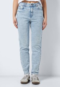 Asmuo, vilkintis šviesiai mėlynus aukštai talpinius džinsus ir kreminės spalvos sportinius batelius, stovi prieš paprastą pilką foną.