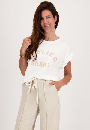Lachende vrouw met lang bruin haar draagt een witte t-shirt met opgerolde mouwen en een beige broek met trekkoord, poseert met één hand in haar zak.