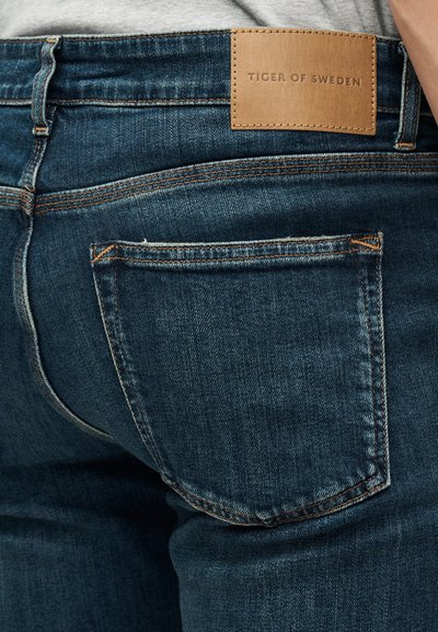 Σκούρα μπλε τζιν με εμφανείς ραφές πίσω τσέπης και καφέ δερμάτινο ταμπελάκι με την επιγραφή «Tiger of Sweden» στη ζώνη της μέσης.