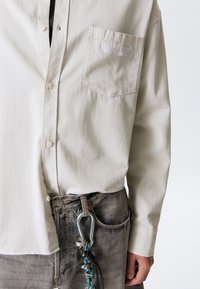 Beige button-up shirt with chest pocket labeled "ATHLETIX DEPT" paired with faded gray jeans and a carabiner keychain hanging from the belt loop.
