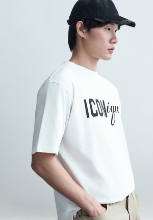 Jeune homme en T-shirt blanc avec le texte "ICON" et pantalon beige, portant une casquette de baseball noire, debout de profil avec les mains dans les poches.
