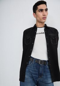 Fekete farmering két mellzsebbel, fehér T-shirt felett, amelyen az "Atomic" felirat látható. Kék farmernadrág fekete övvel, fém részletekkel.