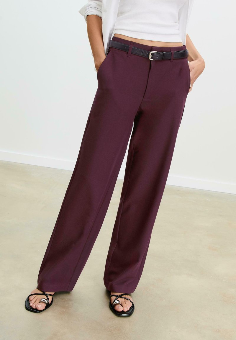 Pantalons larges bordeaux foncé avec une texture subtile, dotés de poches avant et d'une ceinture noire. Associés à des sandales noires à lanières.