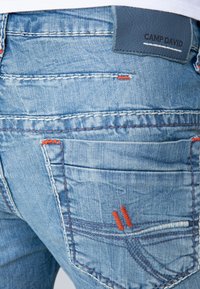 Jeans de mezclilla azul claro con un acabado texturizado, costuras decorativas, acentos en naranja y un parche de cuero de CAMP DAVID en la cinturilla.