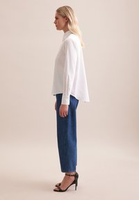 Biała bluzka z długim rękawem i wysokim kołnierzem, zestawiona z niebieskimi dżinsami o prostym kroju z wysokim stanem oraz czarnymi sandałami na wysokim obcasie.