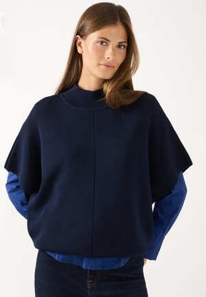 Frau mit langen braunen Haaren trägt einen dunkelblauen, locker sitzenden Kurzarmpullover über einem hellblauen Langarmshirt und dunklen Jeans vor weißem Hintergrund.