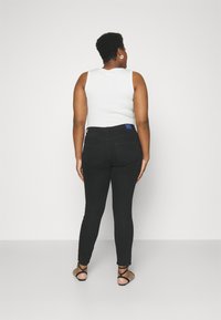 Jeans negros de talle alto con cinco bolsillos y una etiqueta de marca azul, combinados con una camiseta sin mangas blanca y sandalias negras con tiras.