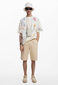 Camicia bianca a maniche corte con motivi astratti colorati, pantaloni corti beige, scarpe beige, cappello a secchiello bianco e occhiali da sole con una borsa a tracolla grigia.
