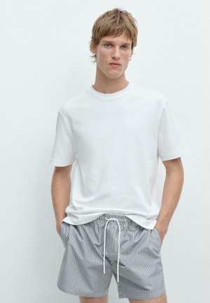 Jonge man die een effen wit T-shirt en marineblauwe en witte verticale gestreepte short met trekkoord draagt, staand met handen in de zakken.