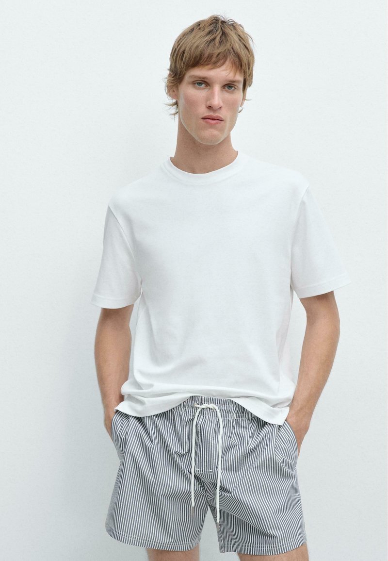 Junger Mann, der ein schlichtes weißes T-Shirt und dunkelblaue und weiße, vertikal gestreifte Shorts mit Kordelzug trägt, steht mit den Händen in den Taschen.