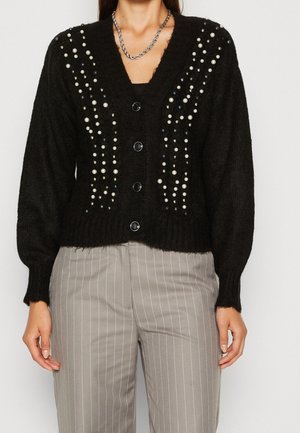 Cardigan noir en maille avec ornements en perles et perles, boutonné sur le devant, porté avec un pantalon beige à fines rayures et un collier chaîne en argent.