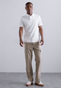Hvit polo-skjorte med krage og svarte knapper, parret med beige cargo-bukser med sidelommer og sneakers med gummisåler.