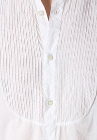 Camisa blanca de botonadura con un diseño texturizado y plisado en la parte delantera superior. Tiene un cuello clásico y botones perlados en color blanco.