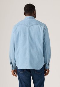 Hele sinine teksasärk terava kraega, pikkade varrukatega ja õmmeldud õlavarre detailiga, kantud tumedate teksapükstega.