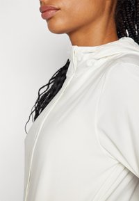 Bijela zip-hoodie s glatkom teksturom, podesivom kapuljačom na vezanje i suptilnim brendiranjem; ima prilagođeni dizajn.