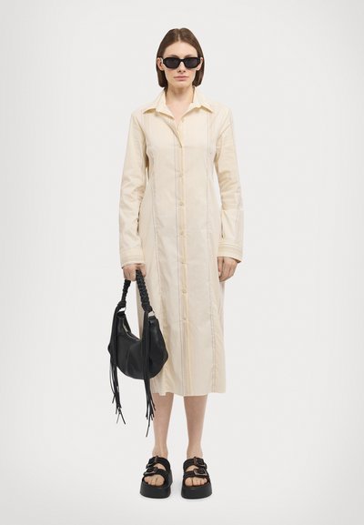 Rochie cămașă bej, cu mânici lungi și cusături verticale. Poartă o geantă neagră și sandale negre cu talpă groasă. Ochelarii de soare negri completează ținuta.
