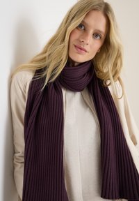 Blonde vrouw met blauwe ogen draagt een geribbelde donkerpaarse sjaal over een beige trui en een lichtbeige jas, leunend tegen een witte wand.