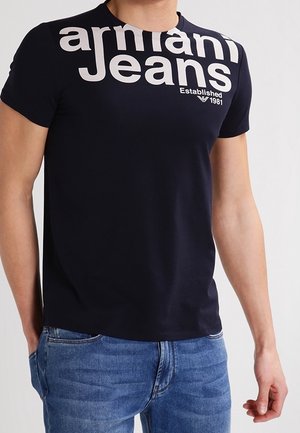 Homme portant un t-shirt noir Armani Jeans avec un logo blanc sur la poitrine et un jean bleu, debout, la main gauche dans la poche.