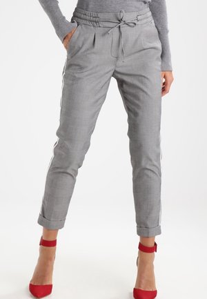 Femme portant un pantalon à carreaux gris avec une taille à cordon et des revers roulés, associé à des talons rouges à bride à la cheville et un haut gris à manches longues.