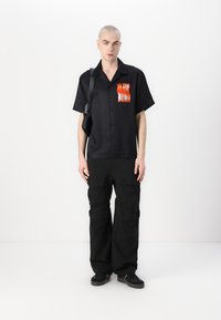 Zwarte kortebloemige shirt met een levendige oranje "SLOW BURN" grafiek, gecombineerd met zwarte cargobroek. De tas heeft een schouderband die kruislings wordt gedragen.