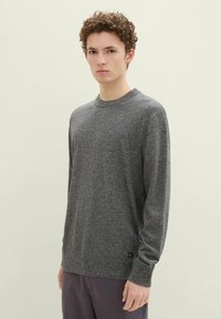 Pull en tricot gris avec un col rond et une finition texturée. Manches longues et détail de logo subtil près de l'ourlet, associé à un pantalon sombre.