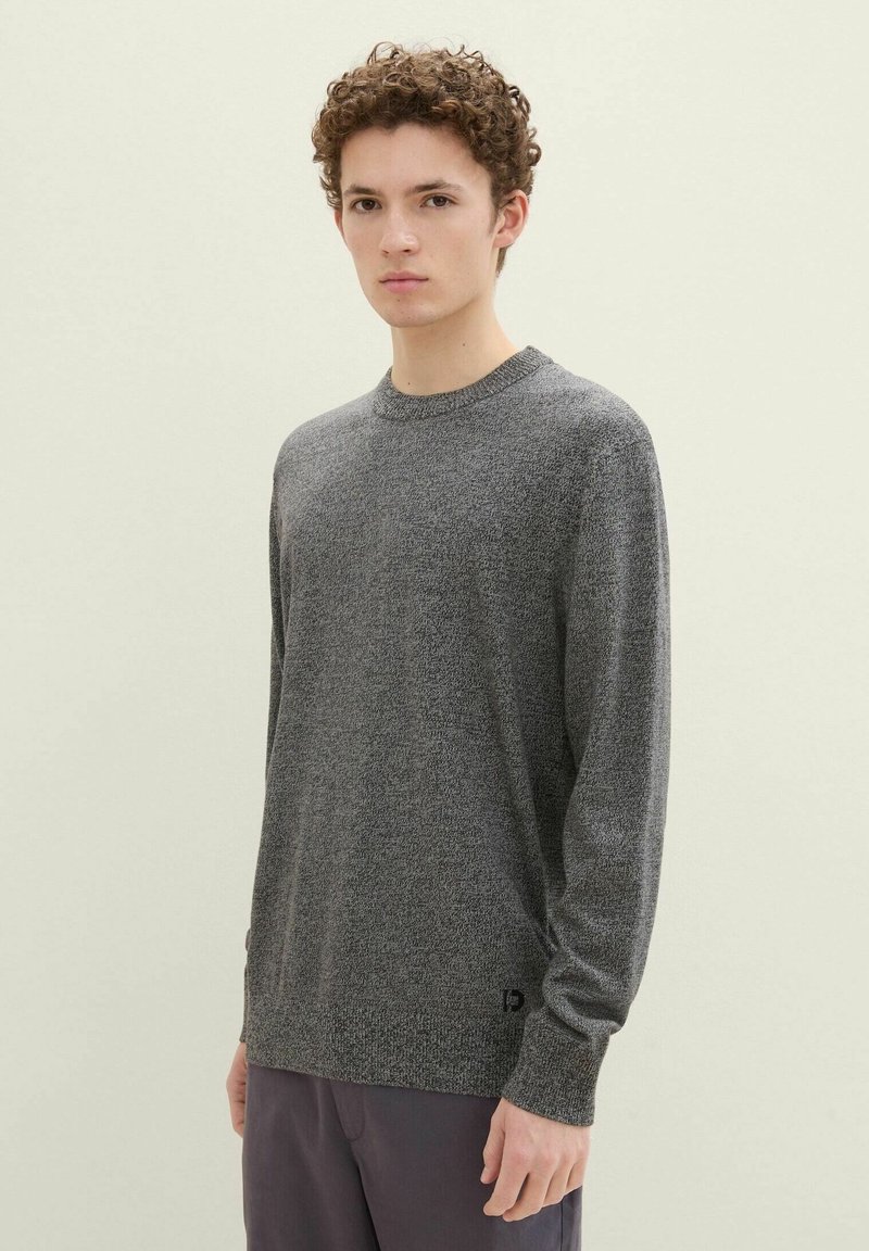 Pull en tricot gris avec un col rond et une finition texturée. Manches longues et détail de logo subtil près de l'ourlet, associé à un pantalon sombre.