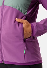 Lila Jacke mit hellgrünem Oberteil, mit einem Reißverschlussfach, weichem Material und elastischen Bündchen. Verfügt über ein Etikett mit grünem Akzent.