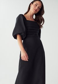 Black dress with a fitted bodice, square neckline, and puff sleeves. The fabric has a smooth texture with horizontal pleats across the chest.