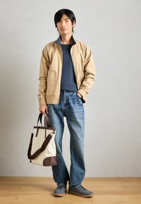 Μπεζ μπομπέρ τζάκετ με μπλε ριγωτό πουλόβερ, ανοιχτό μπλε τζιν και γκρι αθλητικά παπούτσια. Κρατώντας μια καμβά τσάντα ώμου με καφέ λεπτομέρειες.