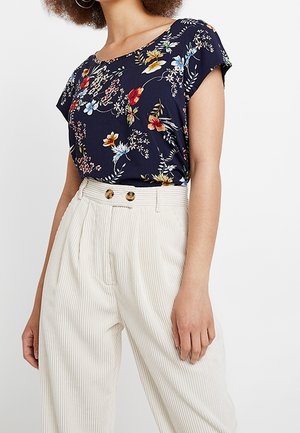 Femme portant un haut à manches courtes bleu marine à fleurs, rentré dans un pantalon large crème taille haute avec deux boutons.