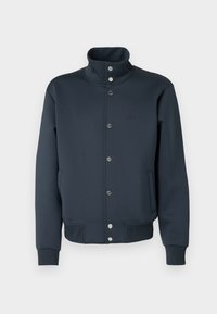 Chaqueta sudadera azul marino con cuello alto, cierre de broches a presión, puños de punto y bolsillos laterales. Cuenta con un logo de Lacoste en el pecho.