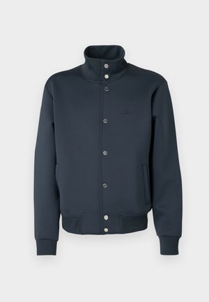 Marineblauwe sweatshirtjas met een hoge kraag, drukknoopsluiting, gebreide manchetten en zijzakken. Voorzien van een Lacoste-logo op de borst.