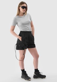 Jeune femme portant un t-shirt gris clair, un short cargo noir, des chaussures de randonnée noires et de grandes lunettes de soleil foncées, debout les mains dans les poches.