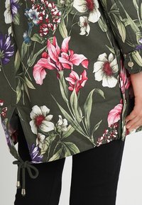Veste en tissu vert olive imprimé floral avec des fleurs roses, blanches et violettes ainsi que des feuilles vert foncé ; comprend un cordon de serrage et des boutons.