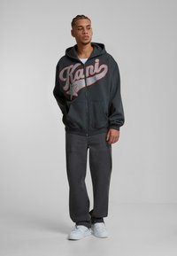 Grå zip-up hoodie med rød og sølv logo, parret med mørke bukser og hvide sneakers. Glat stof og afslappet pasform.