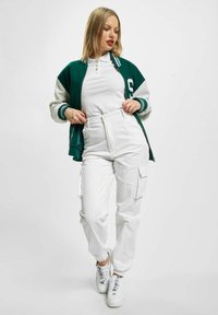 Grønn og hvit bomberjakke med ribbestruktur, over en hvit ribbet topp. Hvite høytlivs cargo-bukser med sidelommer. Hvite joggesko.