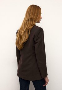 Rjavi blazer z navpičnimi črtami, oblikovanimi ramenih in zareznim reverjem. Material je videti gladek. Stilizirano s temnimi kavbojkami in dolgimi, valovitimi lasmi.