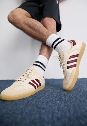 Beige sneakers with maroon stripes, textured upper, and gum rubber sole, paired with ribbed white socks featuring black stripes.
