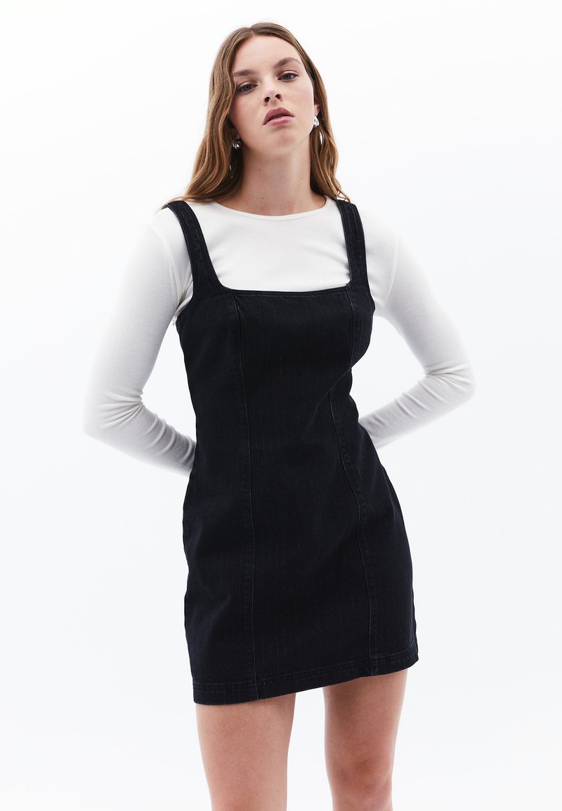 Schwarzes Denim-Kleid mit verstellbaren Trägern, rechteckigem Ausschnitt und figurbetonter Silhouette. Darunter ist ein weißes Langarmoberteil.