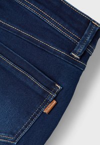 Tumši zilas džinsu bikses ar redzamu dūri, ādas zīmolu virkni un klasisku piecu kabatu dizainu. Gluda tekstūra un izturīgs audums.