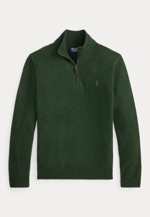 Mann trägt dunkelgrünen Zip-up-Pullover, hellblaues Hemd, dunkelgraue plissierte Hose und braune Tassel-Loafer, steht mit den Händen in den Taschen.