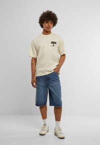 Beige kortærmet t-shirt med "BEWARE" grafisk lomme-detalje, parret med blå denim shorts. Let, afslappet stof med en løs pasform.