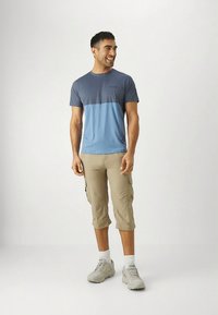 Blå och grå kortärmad T-shirt, beige cargoshorts, ljusgrå sneakers, stående pose, leende mot en neutral bakgrund.
