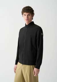 Zwarte zip-up jas met een hoge kraag, voorzien van elastische manchetten, zijzakken en een circulair logo op de linkermouw. Glad materiaal.