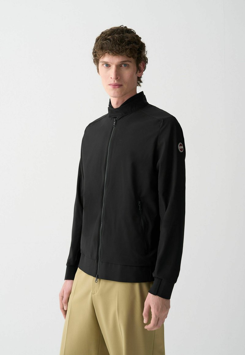 Zwarte zip-up jas met een hoge kraag, voorzien van elastische manchetten, zijzakken en een circulair logo op de linkermouw. Glad materiaal.