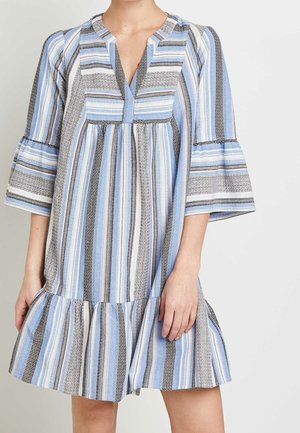 Femme portant une robe ample arrivant aux genoux avec des rayures verticales bleues, blanches et noires, et des manches larges 3/4 avec un ourlet à volants.